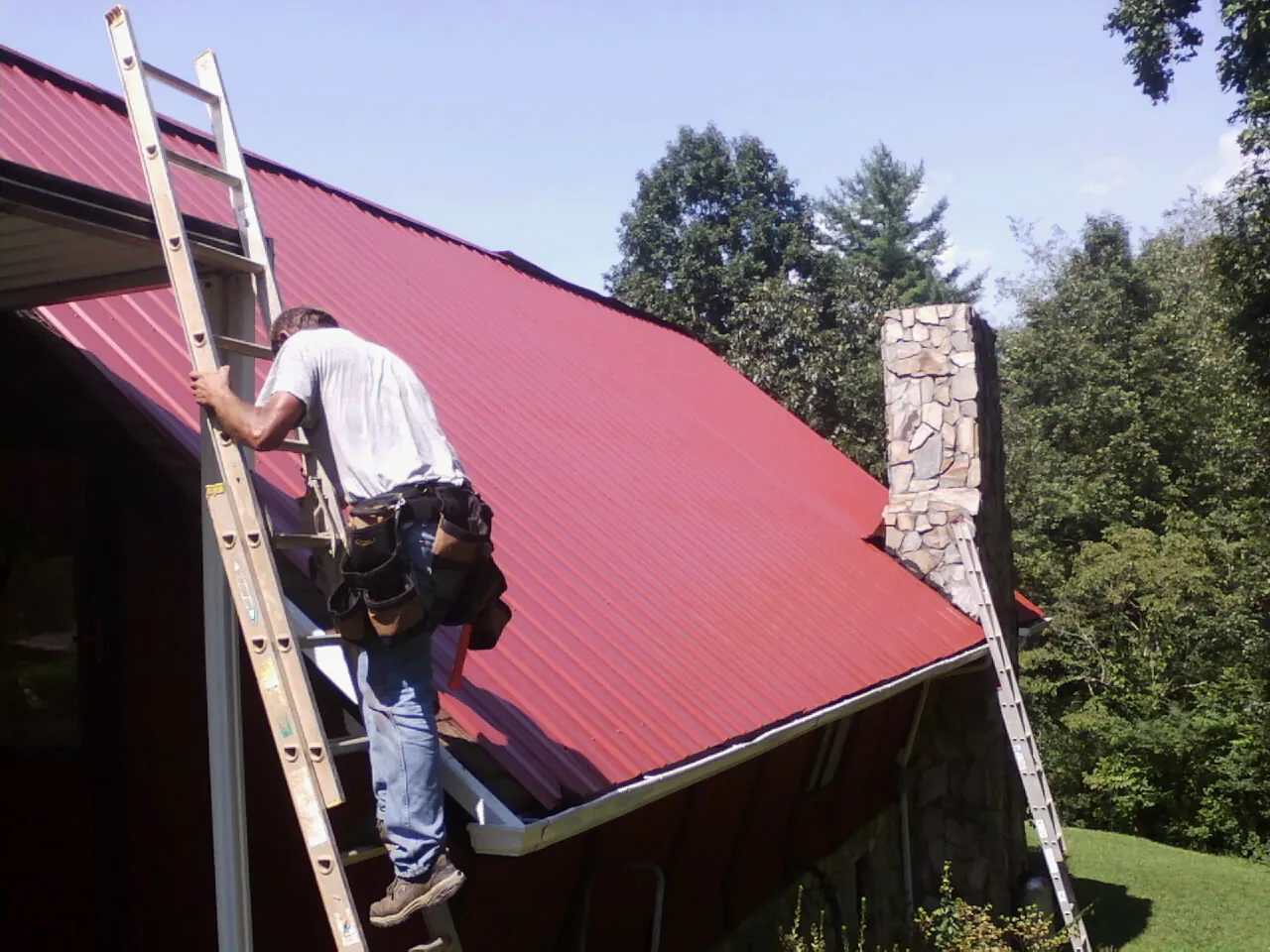Roofer on a ladder beside a red metal roof and stone chimney — Soffit & Fascia Repair by Roofing Brothers in Cave Junction